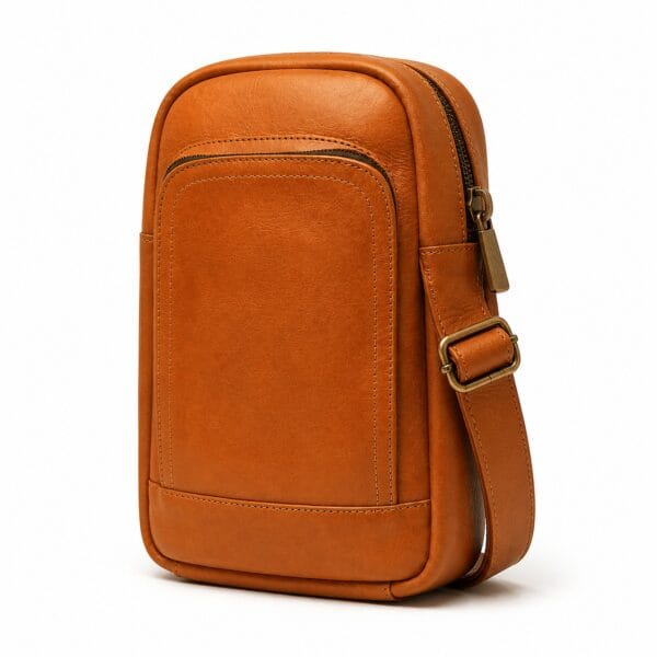 Compact cognac brown full-grain leather sling bag, vertical body with a rounded top, front zip pocket and an adjustable leather strap, shown three-quarter on white.