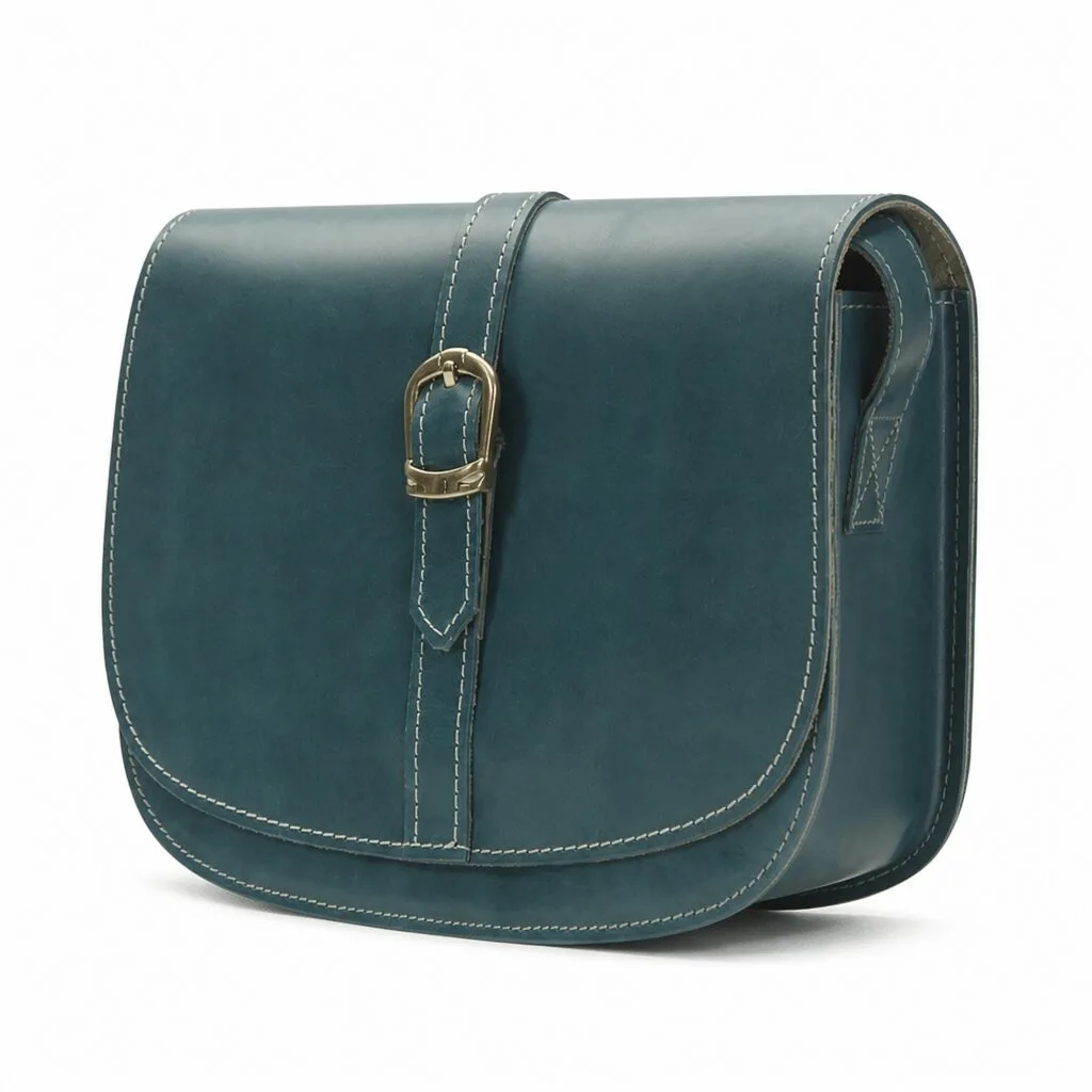 Deep teal leather saddle bag at a three-quarter angle, showing the rounded saddle silhouette, the gold-tone buckle strap and the cream contrast stitching.