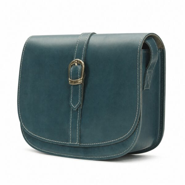 Deep teal leather saddle bag at a three-quarter angle, showing the rounded saddle silhouette, the gold-tone buckle strap and the cream contrast stitching.